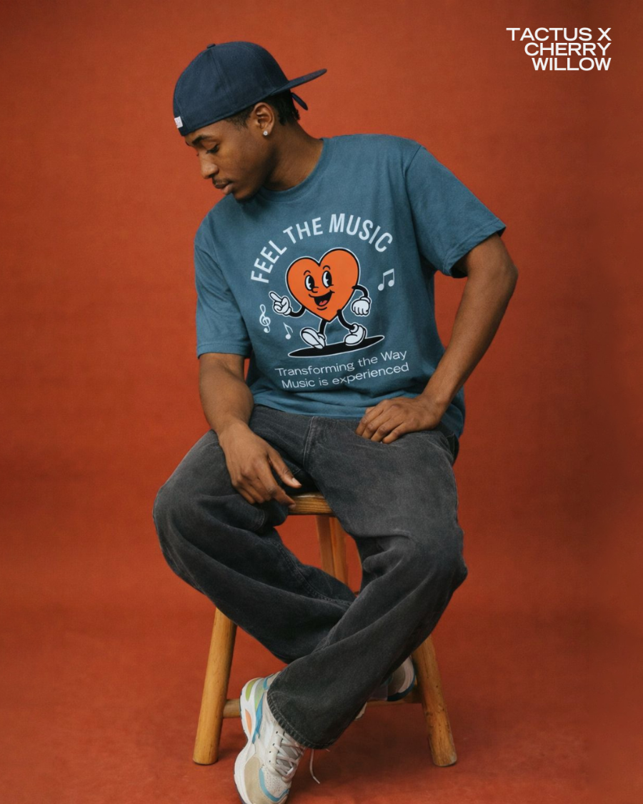 Model seated on a stool wearing the blue CWA × Tactus "Feel the Music" t-shirt against a warm rust backdrop